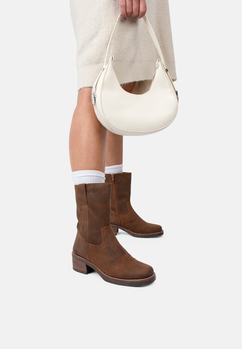 Brown suede ankle boots with a block heel, styled with white socks. Held is a cream-colored crescent-shaped handbag.