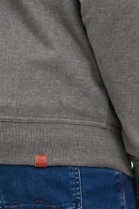 Felpa grigia con polsini e orlo a costine, caratterizzata da un piccolo tag in pelle marrone sulla cucitura inferiore. Indossata sopra dei jeans in denim blu.