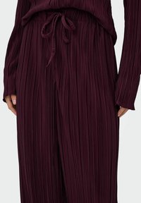 Pantalons larges plissés bordeaux avec un waistband à cordon de serrage, présentant une texture verticale et des manches longues, mettant en valeur un tissu doux et fluide.