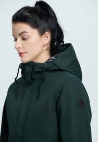 Chaqueta acolchada de color verde oscuro con capucha, que cuenta con cordones ajustables, un diseño elegante y un logotipo negro redondo en el hombro.