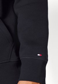 Black sweatshirt with a smooth texture. Features a logo patch in red, white, and blue at the cuff. Stitching is visible along the seam.
