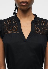 Gros plan d'une personne portant un chemisier noir avec des découpes en dentelle aux épaules et un col en V à volants.