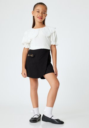 Camisa blanca con cuello bordado, falda envolvente negra con un bolsillo lateral y accesorios dorados, combinada con zapatos negros y calcetas blancas.