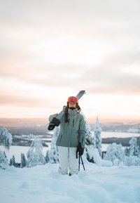Skidutrustning: en kvinna i en ljusgrön jacka, vita byxor och svarta handskar står i snön och håller skidor över axeln.