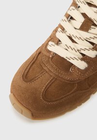 Brown suede sneaker with textured surface, detailed stitching, and cream laces featuring subtle patterns. Slightly rounded rubber sole.