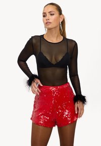 Haut noir transparent à manches longues avec un soutien-gorge bralette en dentelle en dessous, associé à un short rouge pailleté. Accentué par des bracelets en plumes noires.