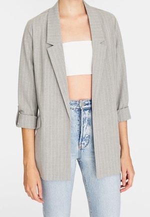 Femme portant un blazer gris clair à fines rayures avec les manches retroussées, un top tube blanc et un jean taille haute bleu clair, debout devant un fond blanc.
