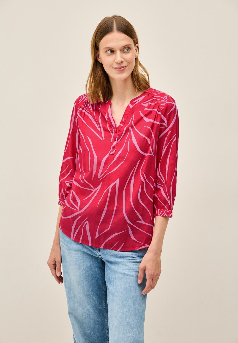 Rote Bluse mit weißem Blattmuster, V-Ausschnitt, Dreiviertelärmel und gerafften Schultern; leichtes Gewebe. Getragen mit hellblauen Jeans.