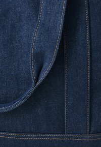 Tumši zils džinsu audums ar redzamām diega šuvēm kontrastējošā oranžā krāsā, ar gludu tekstūru un sānu kabatu.