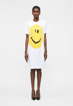 Frau trägt ein weißes knielanges Kleid mit großem gelbem Smiley-Gesicht, steht vor einem schlichten weißen Hintergrund in schwarzen Absätzen.