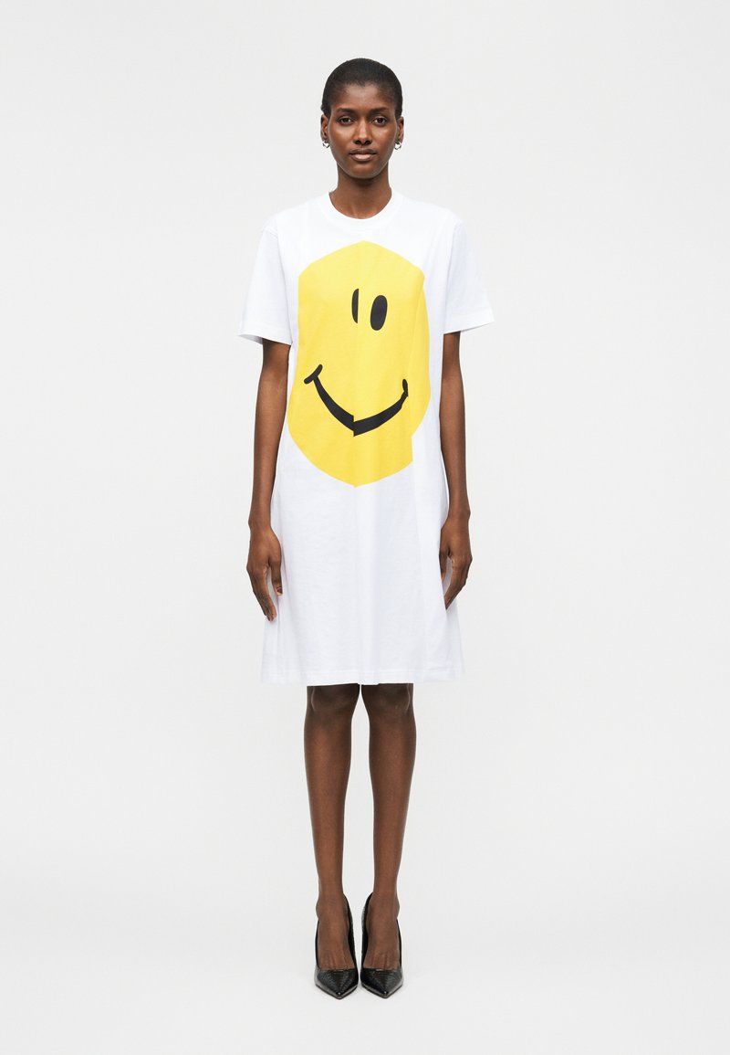 Femme portant une robe blanche mi-longue avec un grand visage souriant jaune, debout devant un fond blanc uni avec des talons noirs.