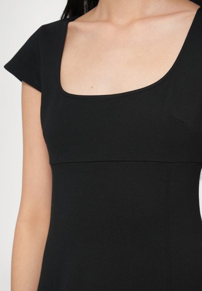 Melna kleita ar kvadrātveida izgriezumu, īsām piedurknēm un gludu audumu. Acīmredzamas šuves uz krūšu daļas uzlabo tās strukturēto formu.