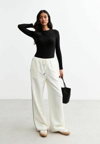 Top negro de manga larga con relieve, combinado con pantalones anchos blancos que presentan franjas laterales negras. Los accesorios incluyen un pequeño bolso negro y zapatillas beige.
