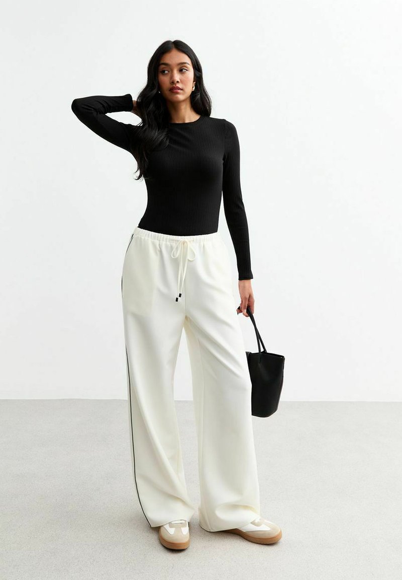 Top negro de manga larga con relieve, combinado con pantalones anchos blancos que presentan franjas laterales negras. Los accesorios incluyen un pequeño bolso negro y zapatillas beige.