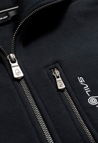 Svart zip-up jacka i mjukt material, med två silvriga dragkedjor och en vit logo på nedersta högra sidan.