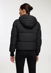 Kobieta z ciemnymi włosami związanymi w kok, ubrana w czarną ocieplaną kurtkę z kapturem, stoi plecami do obiektywu na jednolitym jasnym tle.