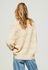 Pull en tricot beige avec motifs géométriques ajourés, coupe décontractée et poignets côtes. Porté avec un jean en denim bleu clair.