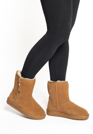 Nohy v černých legínách, obuté v pískových semišových zimních botách s beránkovým zateplením a bočním zipem, stojící s jednou nohou mírně zvednutou.