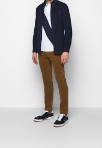 Námořnická košile s knoflíky přes bílý polo triko s diagonálním pruhováním, doplněná hnědými kordurovými kalhotami a černými teniskami s bílými tkaničkami.