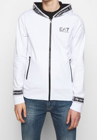 Felpa bianca con zip realizzata in tessuto elasticizzato, con un logo nero sul petto e una zip nera a contrasto. Presenta tasche laterali e polsini con marchio.