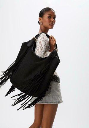 Borsa tote in suede nera con frange lunghe, dotata di doppi manici in pelle e hardware dorato, esposta su uno sfondo neutro.