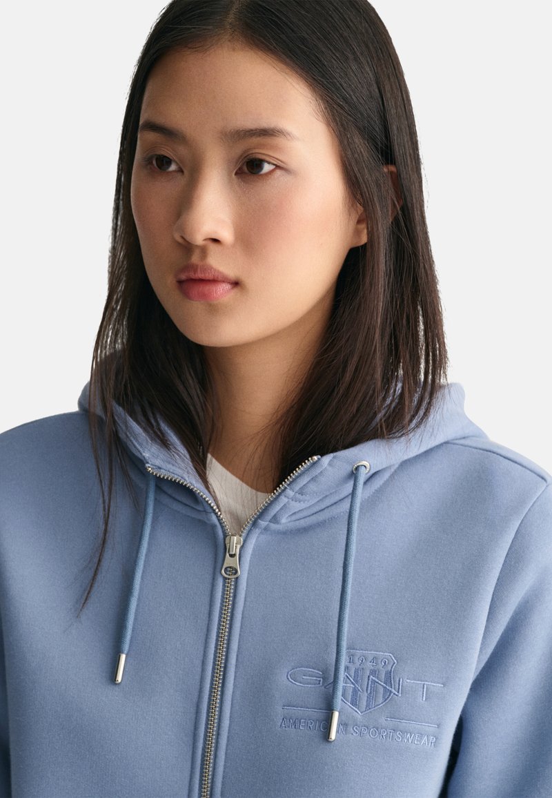 Lichtblauwe zip-up hoodie van zacht materiaal, voorzien van een logo patch aan de voorkant, trekkoorden en zilverkleurige ritshardware.