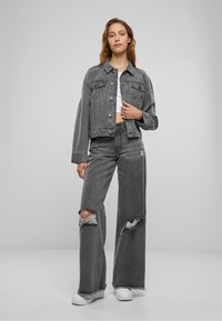 Grå denimjacka och vida jeans med slitna detaljer. Modellen har på sig en vit topp och sneakers. Slät textur och avslappnad passform.