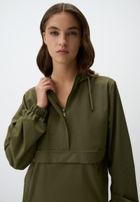 Veste pullover vert olive avec une demi-fermeture éclair, manches élastiques et capuche à cordon de serrage. Texture lisse, design minimaliste, sans motifs visibles.
