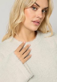 Sweatshirt gris clair, fluffy, avec une finition texturée, un col rond et des manches larges. Gros plan sur une main reposant sur le tissu du vêtement.