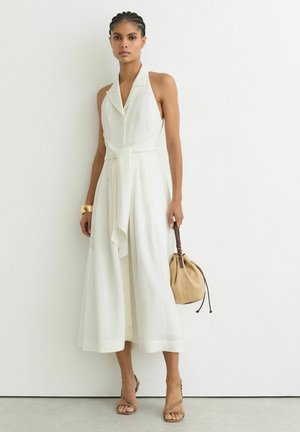 Nainen, jolla on letitetty hius, käyttää hihattoman valkoisen midi-mekon, jossa on solmittu vyötärö, kantaa beigeä säilytyskassia ja käyttää kultaista ranneketjua sekä remmikorkokenkiä.