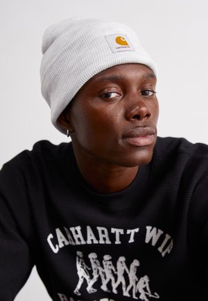 Pelēks ribots beanie ar kvadrātveida Carhartt logo uzlīmi. Melna teksturēta džemperis ar balto grafisko apdruku. Tuva modele sejai.