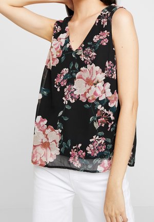 Mouwloos zwart top met V-hals en grote roze-witte bloemenprint, gedragen met witte broek, arm achter het hoofd geheven.