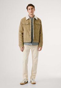 Tan corduroy jack met een witte sherpa kraag, gedragen over een blauw en wit geruite shirt, gecombineerd met crèmekleurige broeken en sneakers.