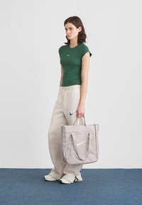 Πράσινο στενό μπλουζάκι, μπεζ φόρμα με λογότυπο και γκρι τσάντα tote με λευκό λογότυπο. Ομαλές υφές, αθλητικός casual σχεδιασμός και ουδέτερα χρώματα.