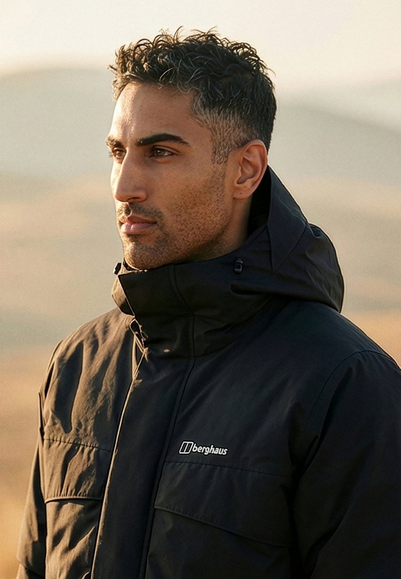 Mand med kort mørkt hår iført sort Berghaus jakke, ser til siden udendørs med sløret naturudsigt i baggrunden.