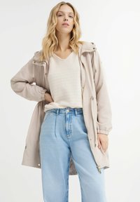 Giacca beige con cappuccio, chiusura con zip, polsini regolabili e vita con cordino, indossata sopra un leggero top di maglia e jeans blu a vita alta.