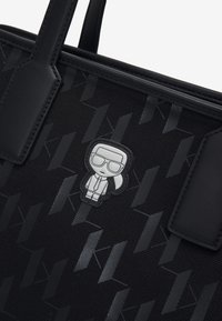 Czarna torba na ramię z teksturowaną powierzchnią z geometrycznym wzorem. Zawiera naszywkę przedstawiającą srebrnego bohatera kreskówkowego z przodu.