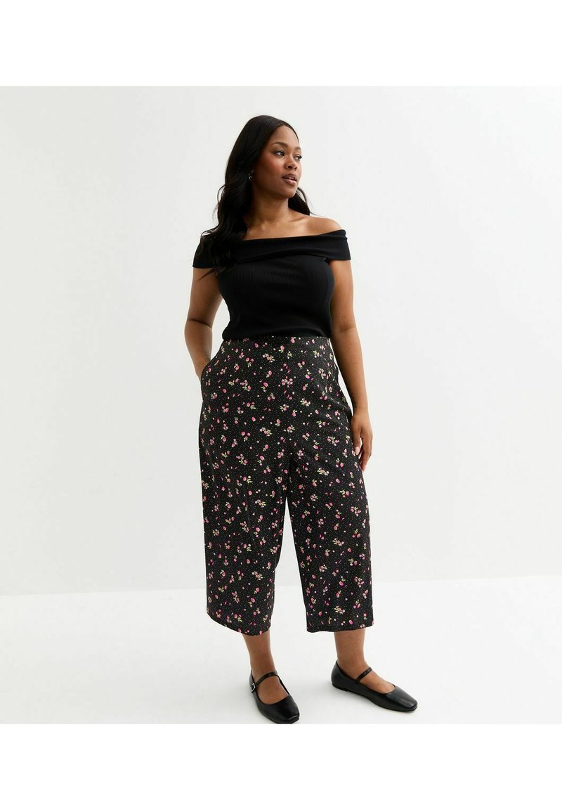 Svart off-the-shoulder ribbad topp som matchar högmidjade, vida byxor i svart tyg med rosa och vita blommönster. Platta svarta skor.