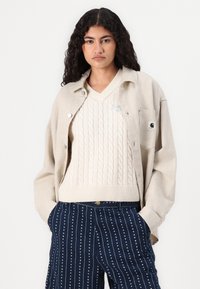 Beige oversized button-up skjorte, kabelstrikket v-hals genser og marineblå stripete bukser med små hvite prikker; uformelle, teksturerte lag.