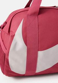 Rosa tygresväska med en stor vit Nike swoosh-logga, texturerat material och robusta remmar. Har meshdetaljer för ventilation.