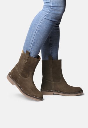 Personne portant un jean skinny bleu clair et des bottines en suède marron avec des languettes, se tenant sur un fond blanc.