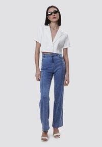 Camisa blanca corta con botones, jeans de mezclilla azul de cintura alta y sandalias de tacón color crema. Los accesorios incluyen gafas de sol negras.