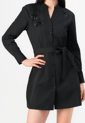 Vestido negro de manga larga con botones, cinturón en la cintura y detalles de cuentas florales en los hombros, usado por una mujer con la mano en la cadera.