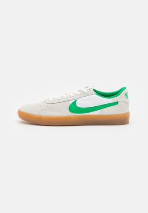 Niski biały sneaker z zielonym logo Nike, zieloną podszewką, białymi sznurowadłami i przezroczystą brązową podeszwą na jednolitym tle.