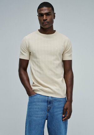 Chemise en tricot beige à manches courtes avec rayures verticales, encolure ras-du-cou côtelée, portée avec un jean en denim bleu, mettant en avant une coupe décontractée et une texture douce.