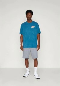 Kék pamut póló rövid ujjakkal és Nike logóval; szürke sportnadrággal és fehér tornacipővel párosítva. Egyszerű, mindennapi sportos öltözék.