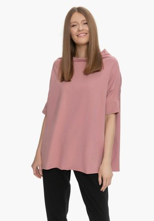 Kobieta z długimi prostymi włosami, uśmiechająca się, ubrana w luźną różową bluzkę z krótkim rękawem i czarne spodnie, stojąca na białym tle.