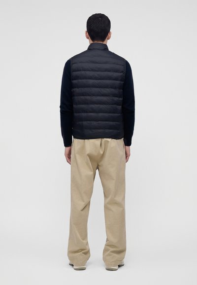 Homme debout de dos, portant un gilet matelassé noir, une chemise noire à manches longues, un pantalon beige et des chaussures claires sur un fond uni.