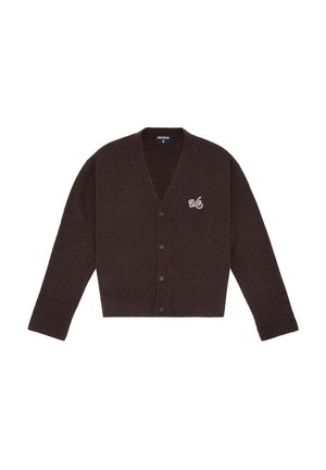 Femme aux cheveux bouclés portant un cardigan marron foncé à boutons, une mini-jupe en cuir noire et des bottes noires montantes jusqu'aux genoux, debout devant un fond gris.