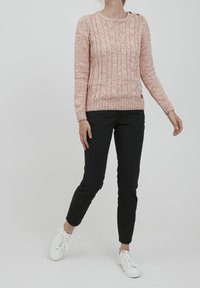 Maglione rosa chiaro a maglia con scollatura rotonda, polsini a coste e dettagli con bottoni. Abbinato a pantaloni neri slim e sneakers bianche.